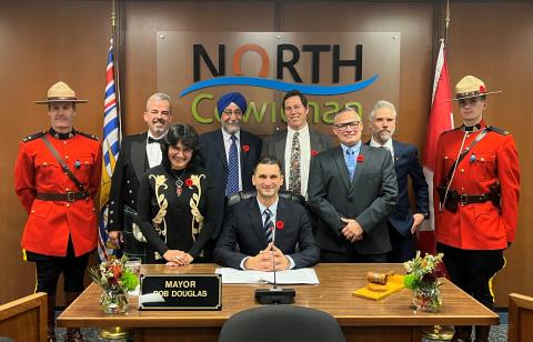 Newly elected Council and Mayor flanked by Mounties. They are all standing behind a large wooden desk.