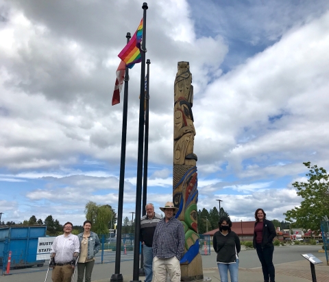 Photo showing people standing below 3 flag pole masts, with the Canadian, B.C., and pride flags